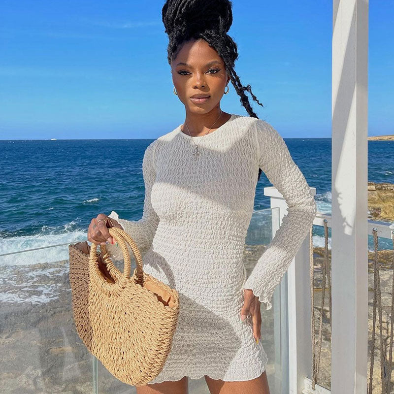 Woman in a white textured long-sleeve mini dress and straw bag, posing with the ocean in the background.