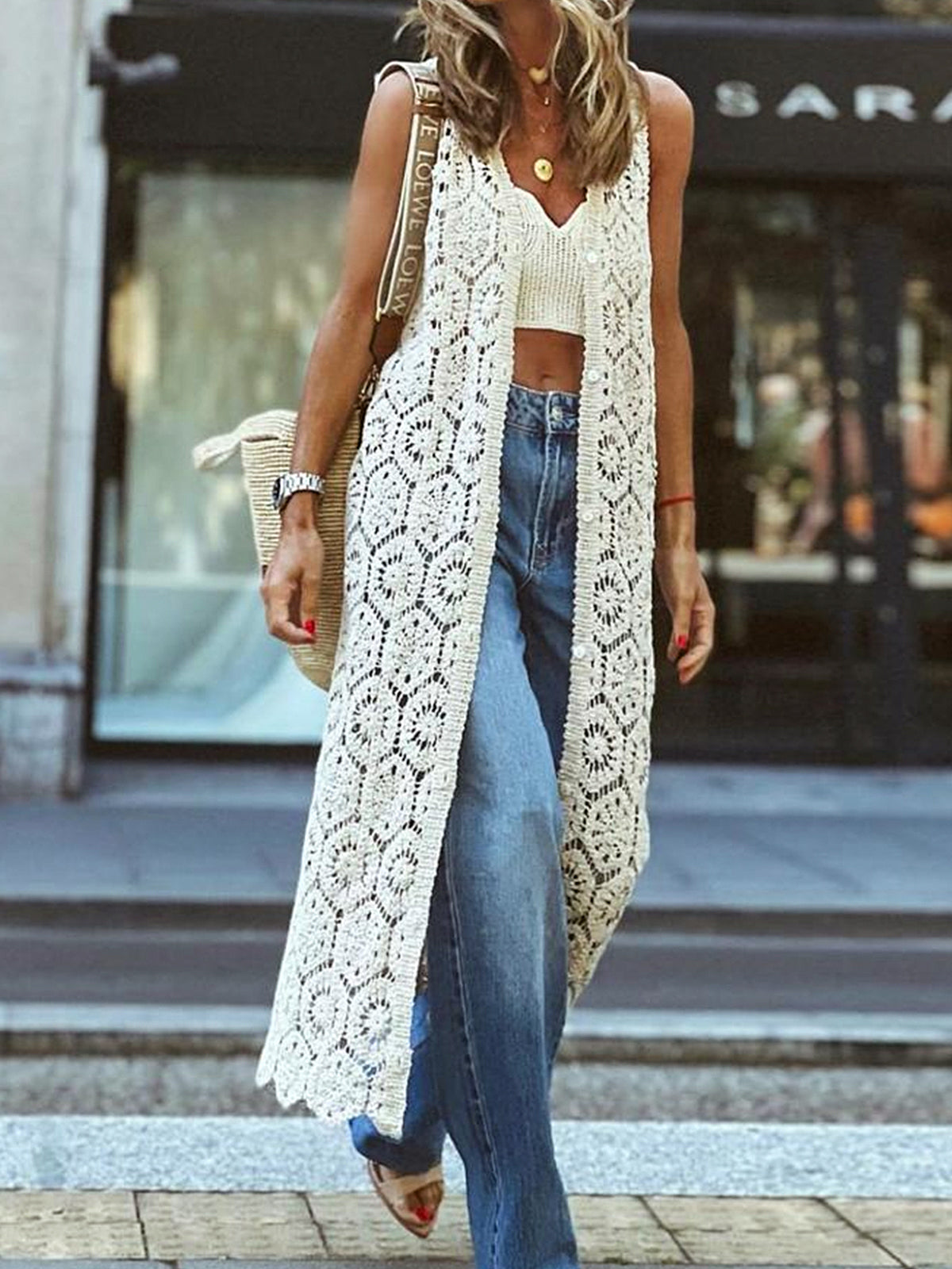 woman wearing a long white crochet vest over a white crop top and blue jeans
