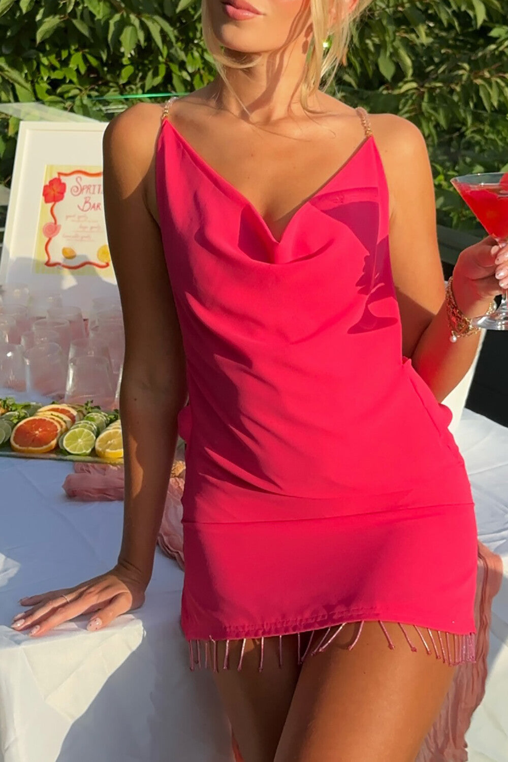 Woman holding a cocktail while wearing a pink mini dress with a beaded fringe hem.