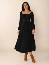 Woman wearing a black midi dress with a square neckline, long puff sleeves, and a corset-style waist detail, standing with her hands at her sides.
