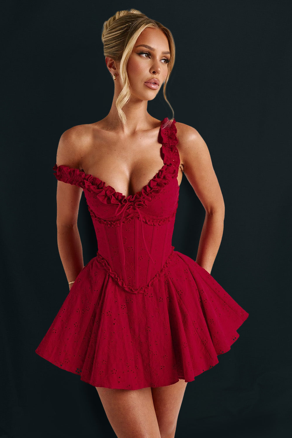 Woman wearing a red off-the-shoulder mini dress with a ruffled neckline and corset bodice, one strap pulled down, against a dark background.