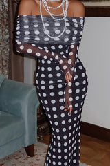 Close-up of a woman's upper body wearing a black and white polka dot off-the-shoulder maxi dress with long sleeves and pearl necklaces, with a light blue couch in the background.