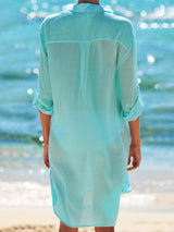Back view of a woman wearing a light blue semi-sheer button-down shirt dress at the beach.
