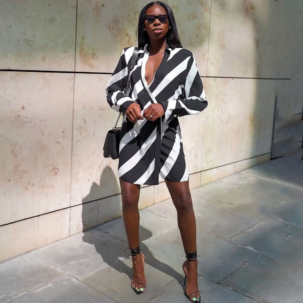 Woman standing and posing in a black and white striped wrap mini dress with long sleeves and a plunging V-neck, accessorized with sunglasses, a black shoulder bag, and black strappy heels.