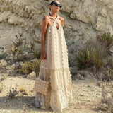 Woman wearing a cream tiered maxi dress with floral appliqué and a halter neckline, standing in a rocky, arid landscape.