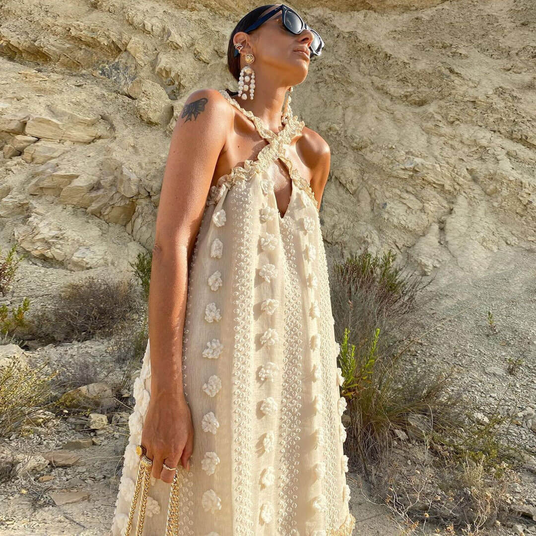 Close-up of a woman wearing a cream tiered maxi dress with floral appliqué and a halter neckline, with a rocky background.