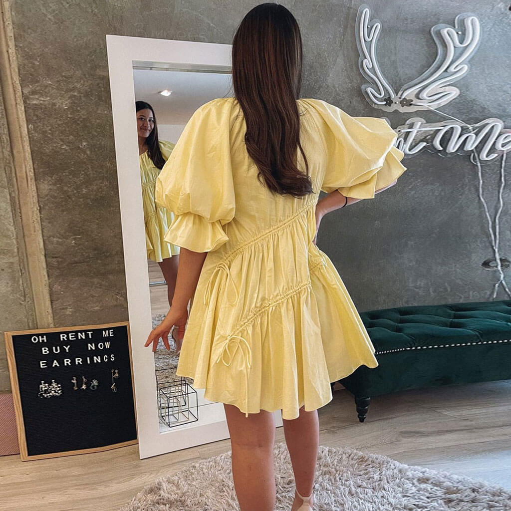 Back view of a woman wearing a yellow mini dress with puff sleeves and drawstring details indoors