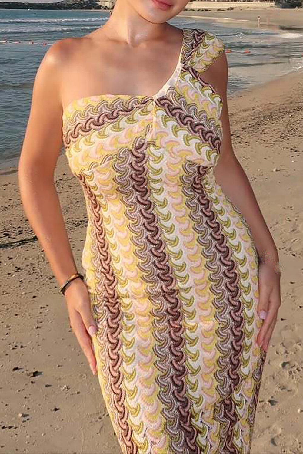 Woman in a patterned one-shoulder dress standing on a sandy beach with the ocean in the background.