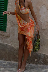 Model wearing an orange and yellow patterned mini dress with draped detailing, holding a textured green bag.