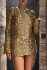 Woman wearing a gold sequined long-sleeved mini dress with a round neckline, standing near wooden paneling.