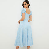 Back view of a woman wearing a light blue floral print midi dress with puff sleeves and a square neckline.