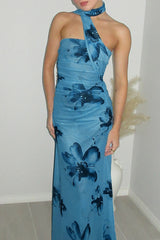 A woman stands in a room wearing a blue floral halter neck maxi dress with draped detailing.