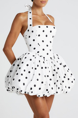 Close-up of a woman wearing a white and black polka dot halter neck dress with a square bodice and a voluminous bubble hem skirt, against a white background.