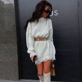 Woman wearing a white oversized knit sweater dress with distressed details, a brown belt, and white boots, standing in front of a black wall.