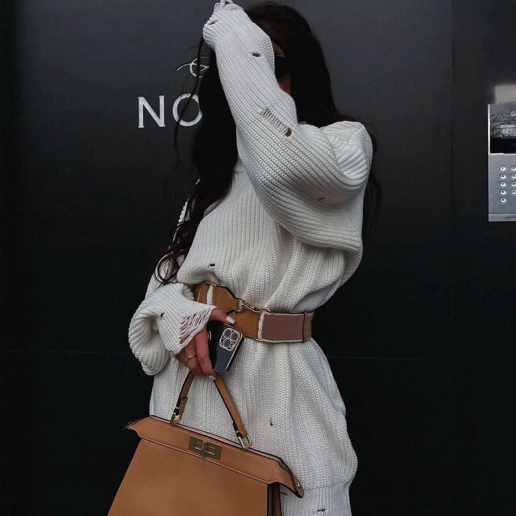 Woman in a white oversized distressed knit sweater dress, accessorized with a brown belt, holding a handbag and a phone.
