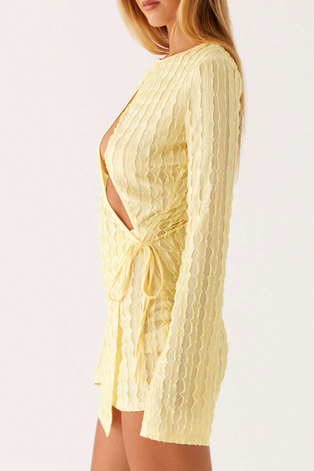 Side view of a woman wearing a light yellow textured mini dress with a plunging neckline and long sleeves, against a white background.
