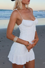 Woman wearing a white textured crinkle mini dress with tie straps and ruffled hem, standing on a beach.