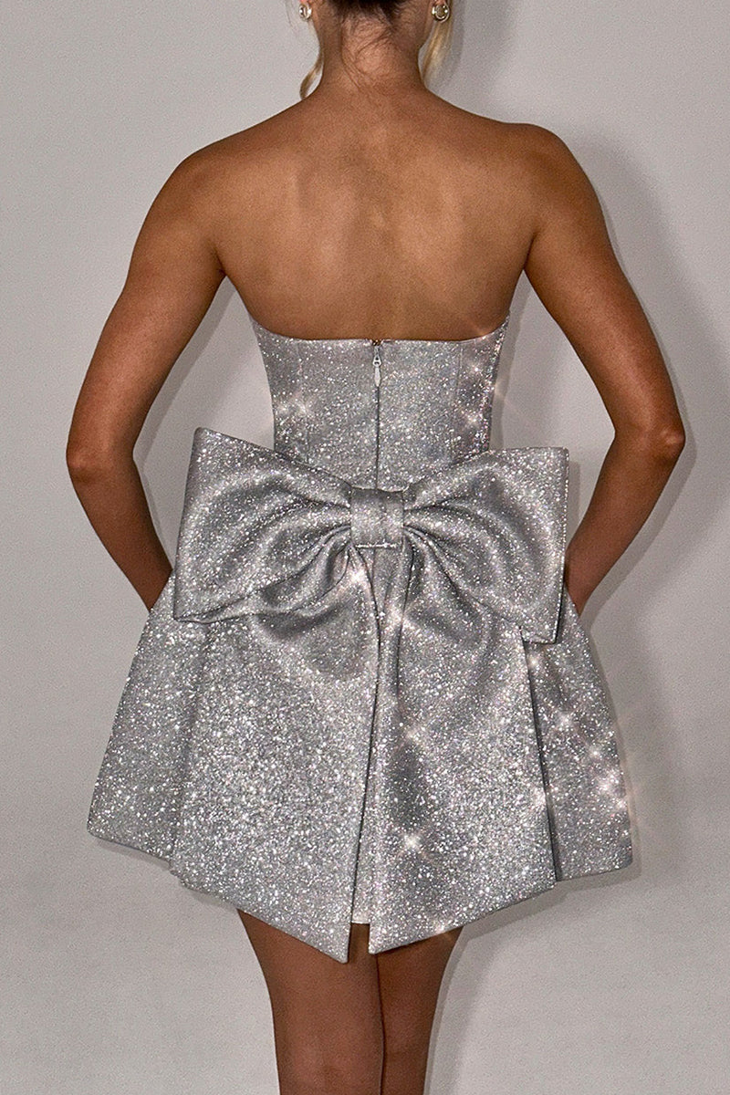 Back view of a woman showcasing a silver sparkly strapless dress with an oversized bow accent on the lower back and a zippered closure.