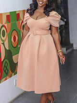 Woman smiling and posing in a light pink off-the-shoulder belted dress with puff sleeves and a pleated midi skirt.
