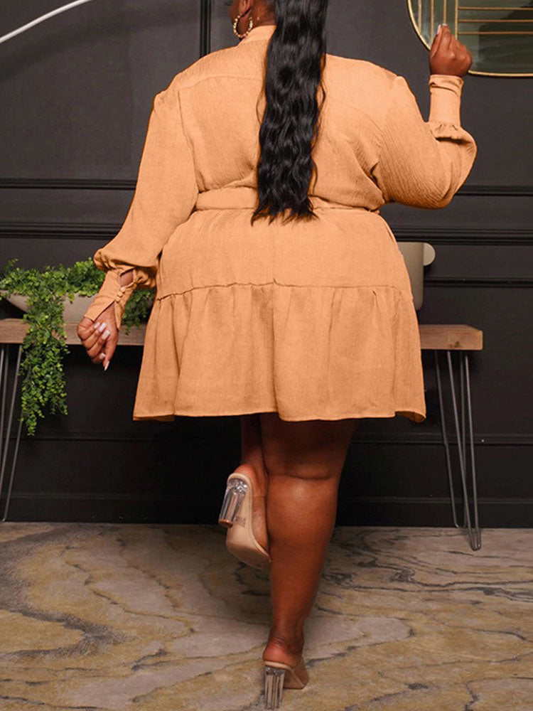 Back view of a plus-size woman wearing a long-sleeved, collared, belted mini dress with a tiered skirt in a peachy-orange color, walking away.