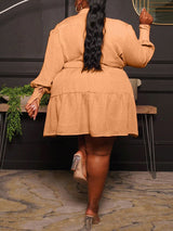 Back view of a plus-size woman wearing a long-sleeved, collared, belted mini dress with a tiered skirt in a peachy-orange color, walking away.