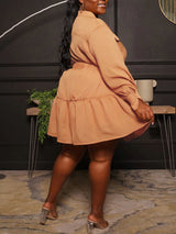 Plus-size woman wearing a long-sleeved, collared, belted mini dress with a tiered skirt in a peachy-orange color, posing with her hand on her hip.