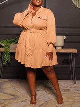 Plus-size woman wearing a long-sleeved, collared, belted mini dress with a tiered skirt in a peachy-orange color.