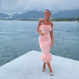woman in a pale pink strapless midi dress with feather trim, standing by the water