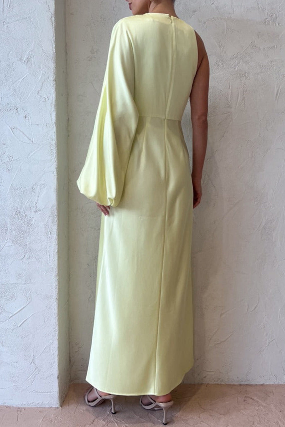 Back view of a woman in a pale yellow satin one-shoulder maxi dress, showing the sleek silhouette and the long asymmetrical sleeve.