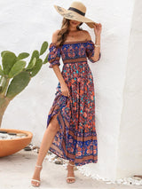 Woman wearing a blue floral off-the-shoulder maxi dress, straw hat, and white sandals, standing next to a cactus.
