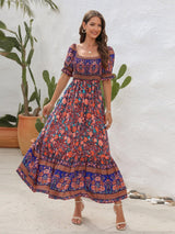 Woman smiling in a blue floral off-the-shoulder maxi dress, with puff sleeves and tiered skirt, wearing white sandals.