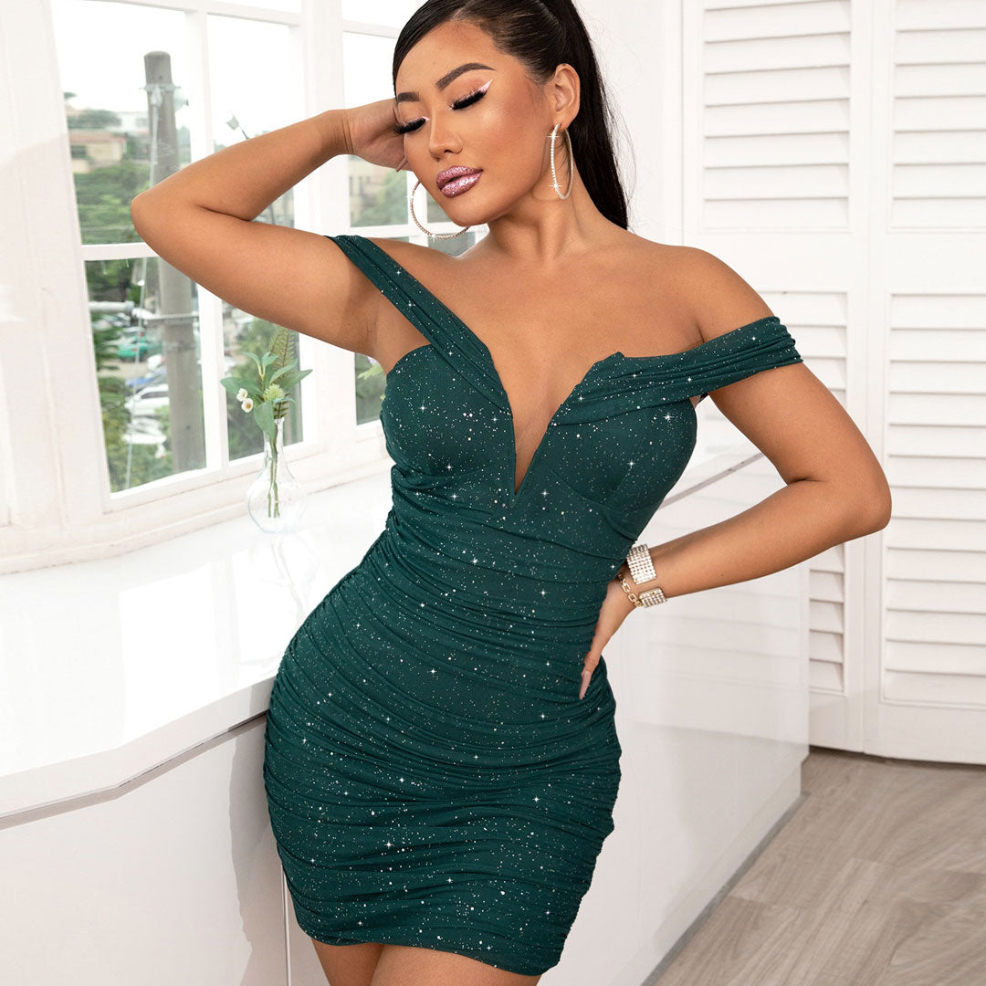 Woman wearing a sparkling green off-the-shoulder ruched bodycon mini dress, posing by a window.