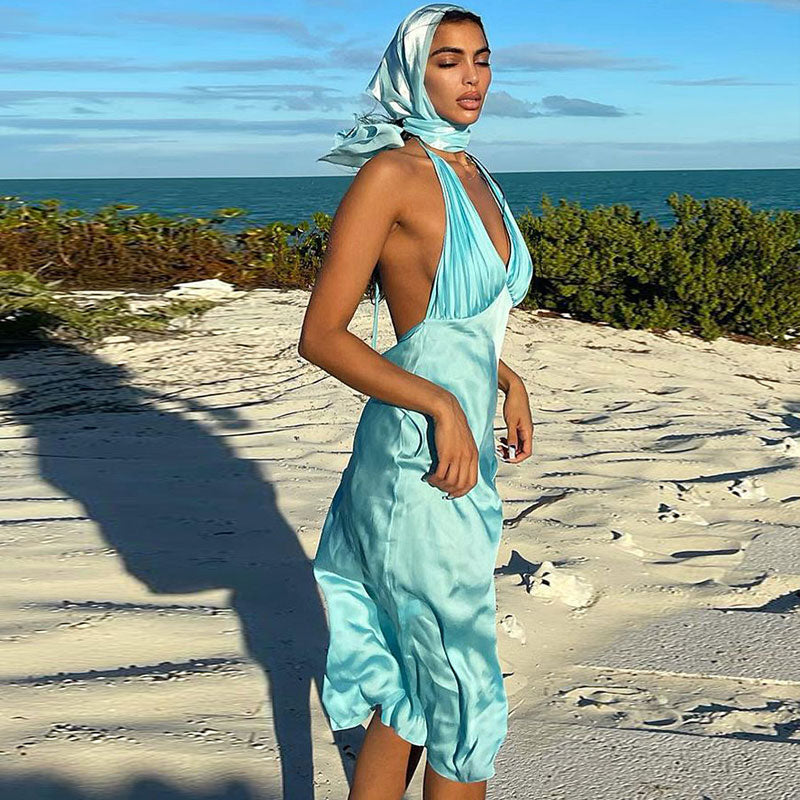 Woman in a light blue satin slip dress with a halter neckline and open back, standing on a sandy beach with the ocean in the background.