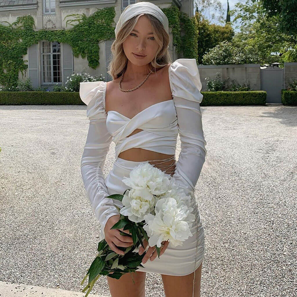 Woman in a white satin dress with long sleeves and a sweetheart neckline, holding a bouquet of white flowers.