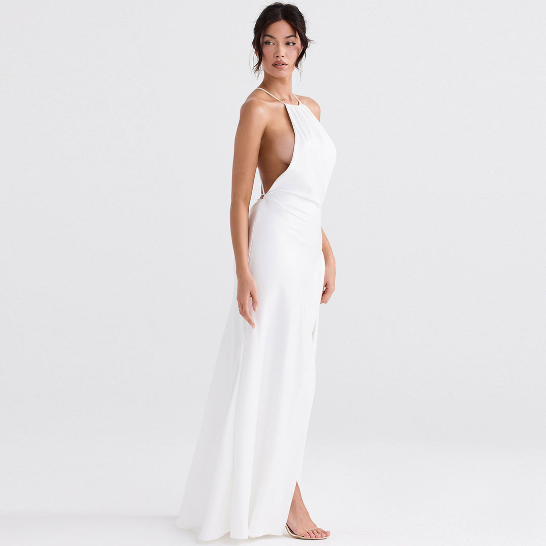 Woman wearing a white satin halter neck maxi dress with a high slit and open back, standing side view