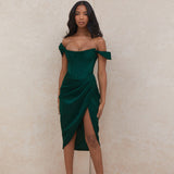 Woman wearing a dark green off-the-shoulder satin dress with a corset bodice and a high slit, standing against a neutral background.