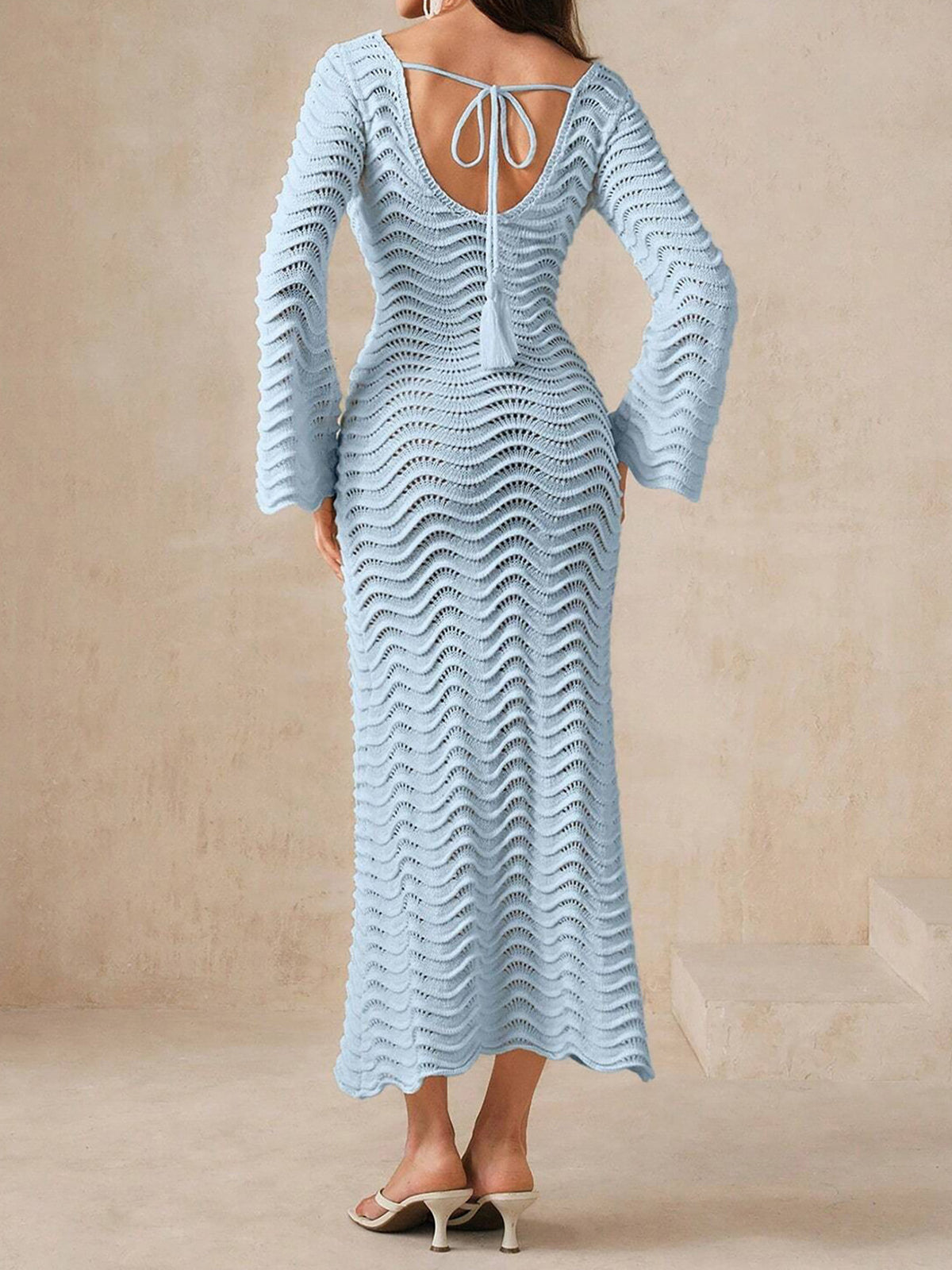 Back view of a woman wearing a light blue, long-sleeved, open-knit maxi dress with a wavy texture and bell sleeves, featuring a tie closure at the back.