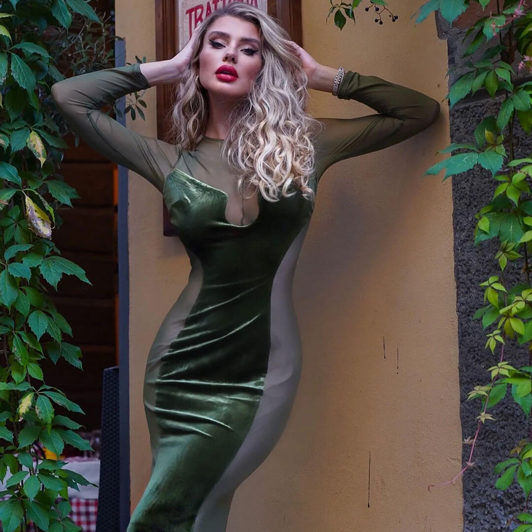 Woman in an olive green velvet and mesh bodycon dress with her arms raised, leaning against a textured wall with ivy