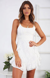 Woman in a white floral jacquard mini dress, standing indoors with a plant and ladder in the background.