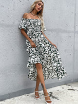 Woman leaning against a wall, wearing a floral print off-the-shoulder maxi dress with a ruffled hem.