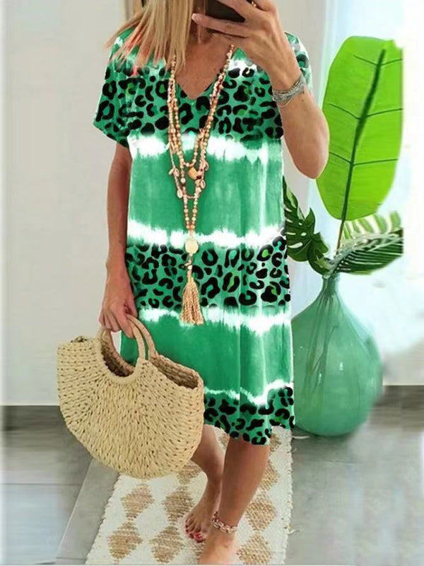 Woman wearing a green tie-dye and leopard print short-sleeve V-neck A-line dress, holding a straw bag, in a room with a plant.