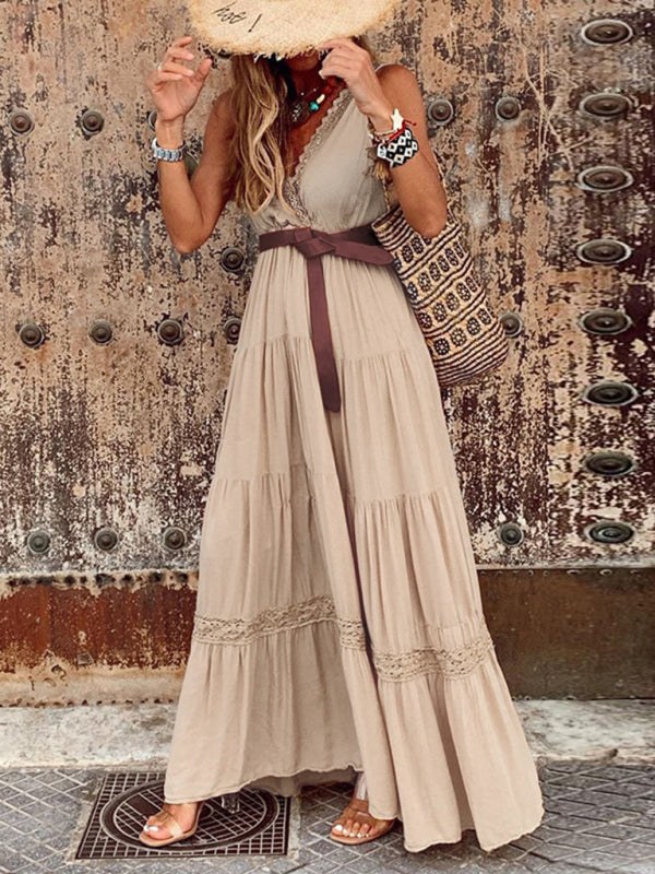 Model showcases a beige sleeveless maxi dress with a V-neck, tiered skirt, and lace detailing, cinched with a brown sash and paired with a straw hat and tote.