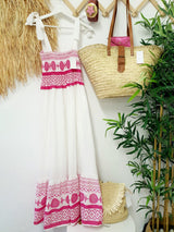 A white and pink embroidered maxi dress hanging on a wall, next to a straw bag and plants.