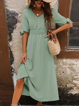 Woman wearing a sage green A-line maxi dress with a smocked waist and tie sleeves, lifting the hem of the dress to reveal her legs.