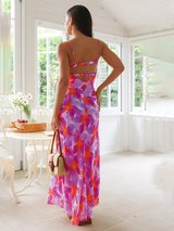 Rear view of a woman wearing a purple and orange floral print maxi dress with spaghetti straps, a ruched bust, and a cut-out back detail.