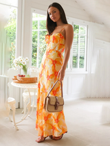 Woman wearing an orange and yellow floral print maxi dress with spaghetti straps and a ruched bust, holding a woven straw handbag.
