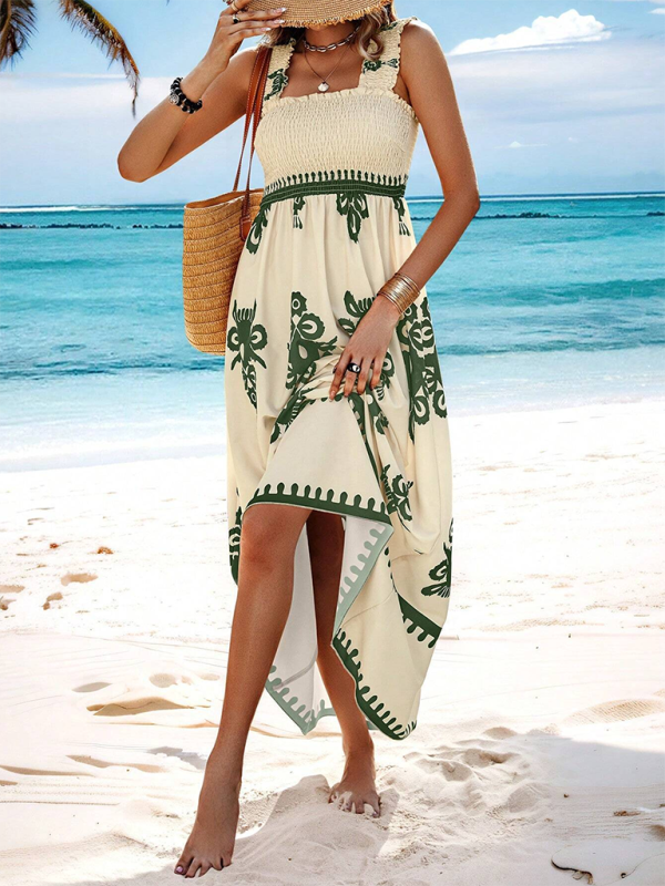 Woman standing on a beach wearing a cream and green floral print maxi dress with smocked bodice and square neckline, holding a straw hat.