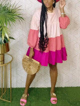 Woman wearing a pink and purple tiered dress, gold chain necklace, gold bracelet, bamboo handbag, and pink sandals, with a hat.