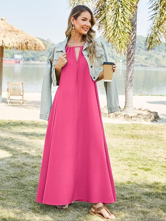 Woman wearing a bright pink sleeveless maxi dress with a halter neckline and keyhole cutout, with a light denim jacket draped over her shoulders, holding a coffee cup, standing on grass by a body of water.