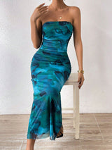 Woman sitting on a stool, wearing a strapless blue and green abstract floral print maxi dress with a high slit.